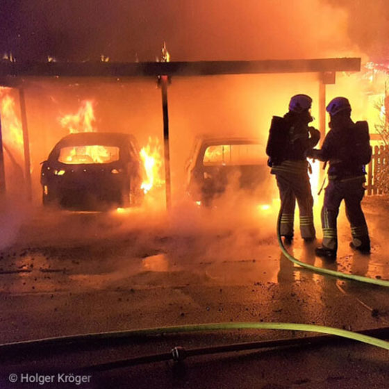 Das Bild zeigt einen dramatischen Einsatz: Zwei Fahrzeuge brennen unter einem Carport, während zwei Feuerwehrleute mit einem Löschschlauch im Schein der Flammen agieren. Die Szene ist vom Licht des Feuers beleuchtet.