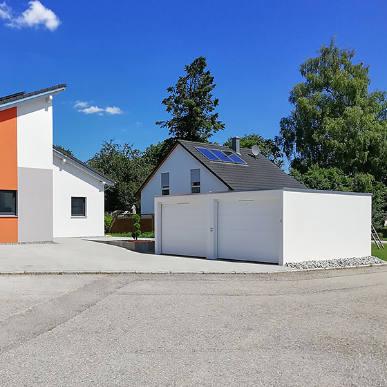 Abgebildet ist eine freistehende Doppelgarage in Weiß mit zwei weißen Garagentoren. Im Hintergrund erkennt man zwei Einfamilienhäuser, die der Szene einen wohnlichen, familienfreundlichen Kontext geben.
