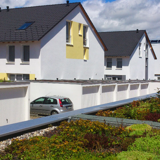 Reihengaragenanlage - Das Bild zeigt Reihengaragen vor mehreren Mehrfamilienhäuser. Die Garagen sind weiss und auf dem Dach sind die mit Moos und Gräsern bewachsen.