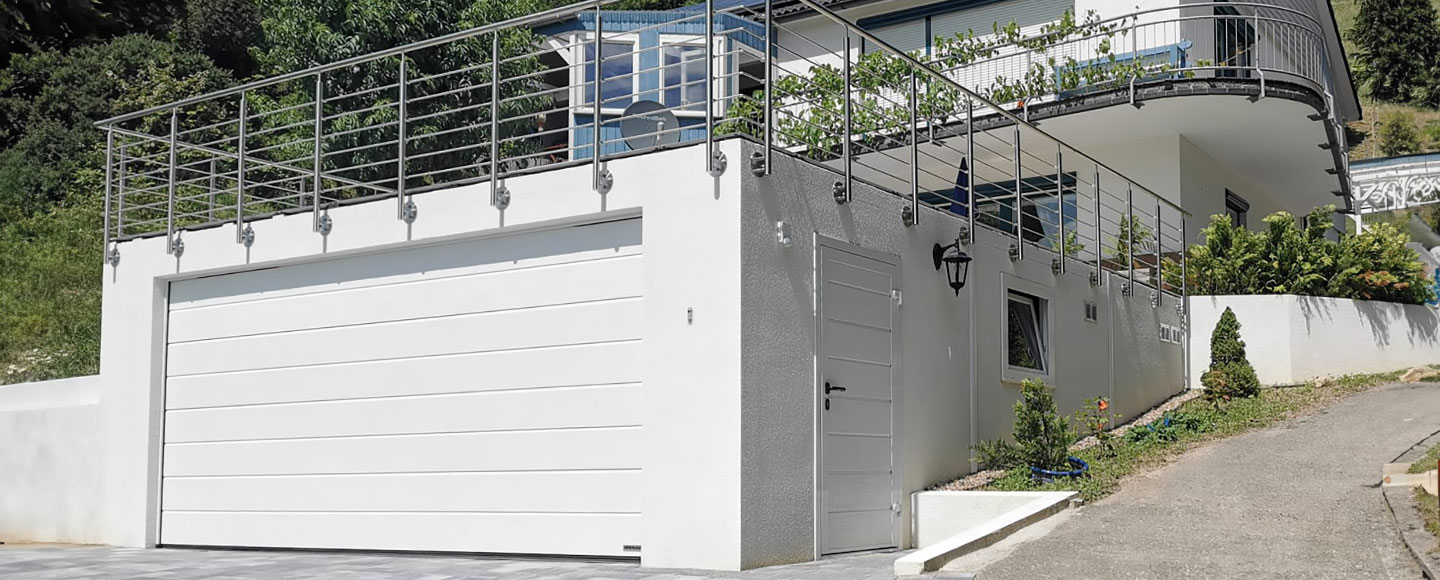 Sie sind auf der Suche nach der perfekten Garage? - Das Bild zeigt eine weiß gestrichene Großraumgarage mit einer Seitentür. Der gepflasterte Hof vor der Garage sorgt für eine aufgeräumte Umgebung. Ein Edelstahlgeländer um das Garagendach weist darauf hin, dass sich dort eine Terrasse befindet. Im Hintergrund ist das dazugehörige Wohnhaus zu sehen.