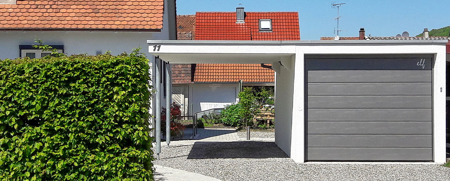 Das Bild zeigt eine moderne Carport-Garagen-Kombination in Weiß. Das Garagentor ist in einem eleganten Grauton gehalten. Der Carport wird von zwei metallisch glänzenden Stützsäulen getragen. Links neben der Garage steht ein gepflegter, grüner Strauch, der das Gesamtbild auflockert und begrünt.