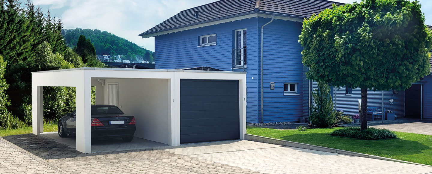 Das Bild zeigt eine moderne weiße Garage mit dunklem Tor, an die ein ebenfalls weißer Carport angebaut ist. Unter dem Carport steht ein schwarzes Mercedes Coupé. Rechts angrenzend befindet sich ein auffälliges blaues Wohnhaus mit Giebeldach, das vollständig mit blau gestrichenem Holz verkleidet ist. Vor dem Hauseingang steht eine farblich passende blaue Bank.