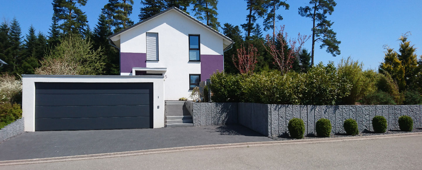 Die Aufnahme zeigt eine weiße Großraumgarage mit dunklem Tor, die sich harmonisch in die Gesamtarchitektur der Wohneinheit integriert. Eine hohe Steinmauer rahmt den erhöht liegenden Rasen und das dahinter befindliche Wohnhaus ein. Über fünf Steinstufen gelangt man hinauf zum Haus, das in den Farben Weiß und Violett gestaltet ist. Vor der Mauer stehen fünf kleine, rund geschnittene Buchsbäume, die für ein gepflegtes Erscheinungsbild sorgen.