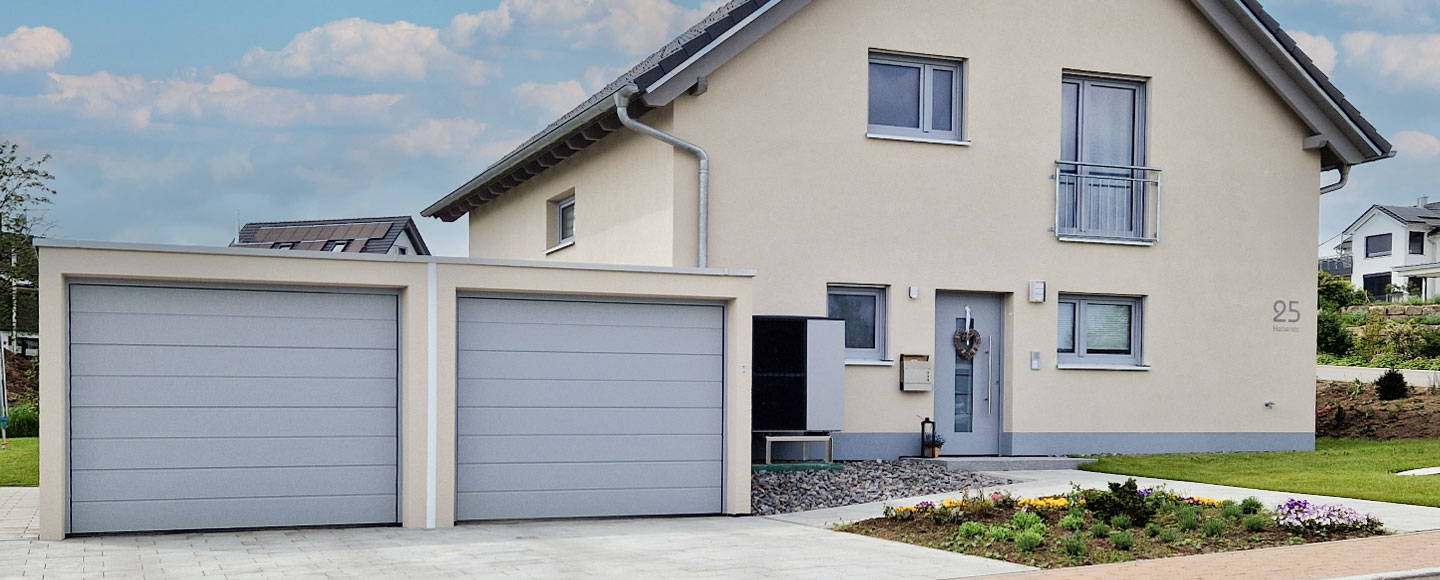 Das Bild zeigt eine beige gestrichene Doppelgarage mit silbergrauen Toren. Rechts und etwas zurückversetzt befindet sich das dazugehörige Familienhaus, das farblich perfekt auf die Garage abgestimmt ist. Auch die Fensterrahmen des Hauses sind in Silber/Grau gehalten und unterstreichen das harmonische Erscheinungsbild von Gebäude und Garage.
