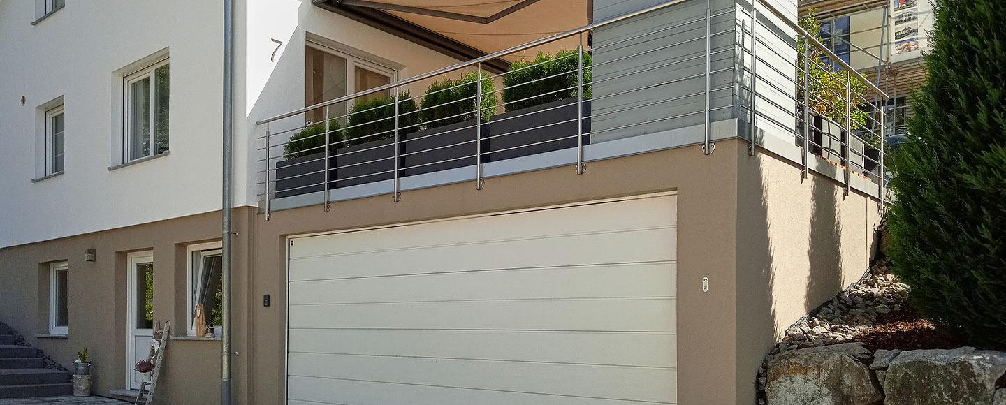 Das Bild zeigt eine braune Großgarage mit weißem Tor, die direkt zwischen einem Wohnhaus und dem Garten eingefasst ist. Das erste Stockwerk des Hauses ist ebenfalls braun gestrichen, während der Rest der Fassade weiß ist. Auf der Garage befindet sich eine Terrasse, die von einem Metallgeländer umgeben ist. Auf der Terrasse stehen zahlreiche schwarze Blumentöpfe mit grünen Büschen, die als Sichtschutz dienen