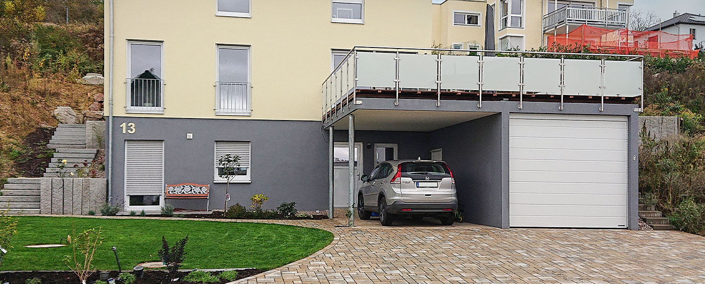 Das Bild zeigt eine dunkelgraue Garagen-Carport-Kombination, die direkt an ein Wohnhaus angebaut ist. Der untere Stock des Hauses ist ebenfalls dunkelgrau, während ab dem zweiten Stockwerk die Fassade hellgelb ist. Die Dachterrasse ist von einem Metallgeländer mit Milchglaseinsatz umgeben. Links am Wohnhaus führt eine Treppe nach oben.