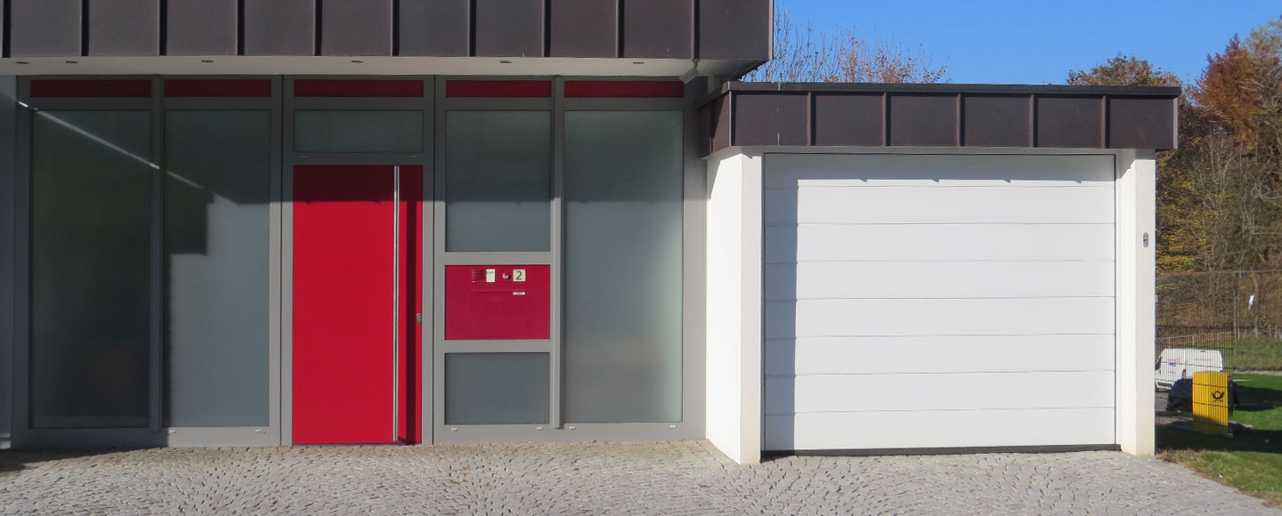 Rechts im Bild befindet sich eine weiße Garage mit passendem weißem Tor. Direkt angebaut ist links ein modernes Flachdachhaus mit auffälliger roter Haustür und rotem Briefkasten. Die restliche Fassade des Hauses besteht aus transluzentem Milchglas, was dem Ensemble ein zeitgemäßes, stilvolles Erscheinungsbild verleiht.