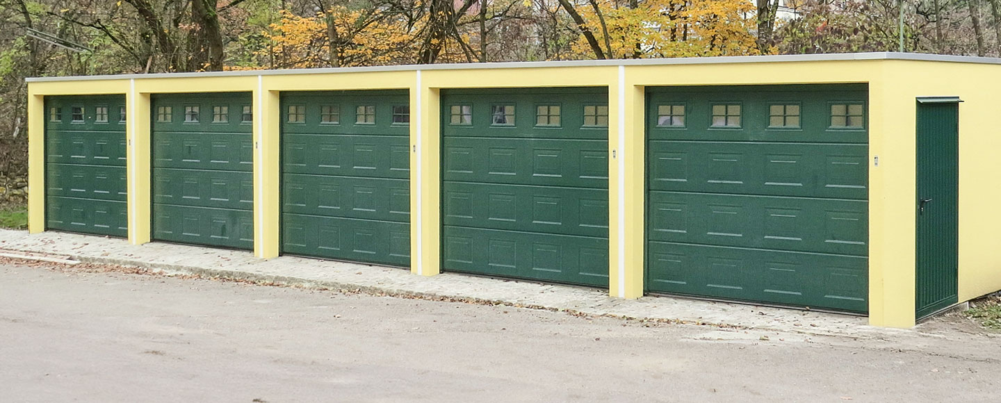 Das Bild zeigt fünf nebeneinander gebaute Garagen, deren Wände gelb gestrichen sind. Die Garagentore sind dunkelgrün und verfügen im oberen Bereich über kleine integrierte Fenster. Rechts an der Seite ist eine dunkelgrüne Tür, die farblich zu den Toren passt.
