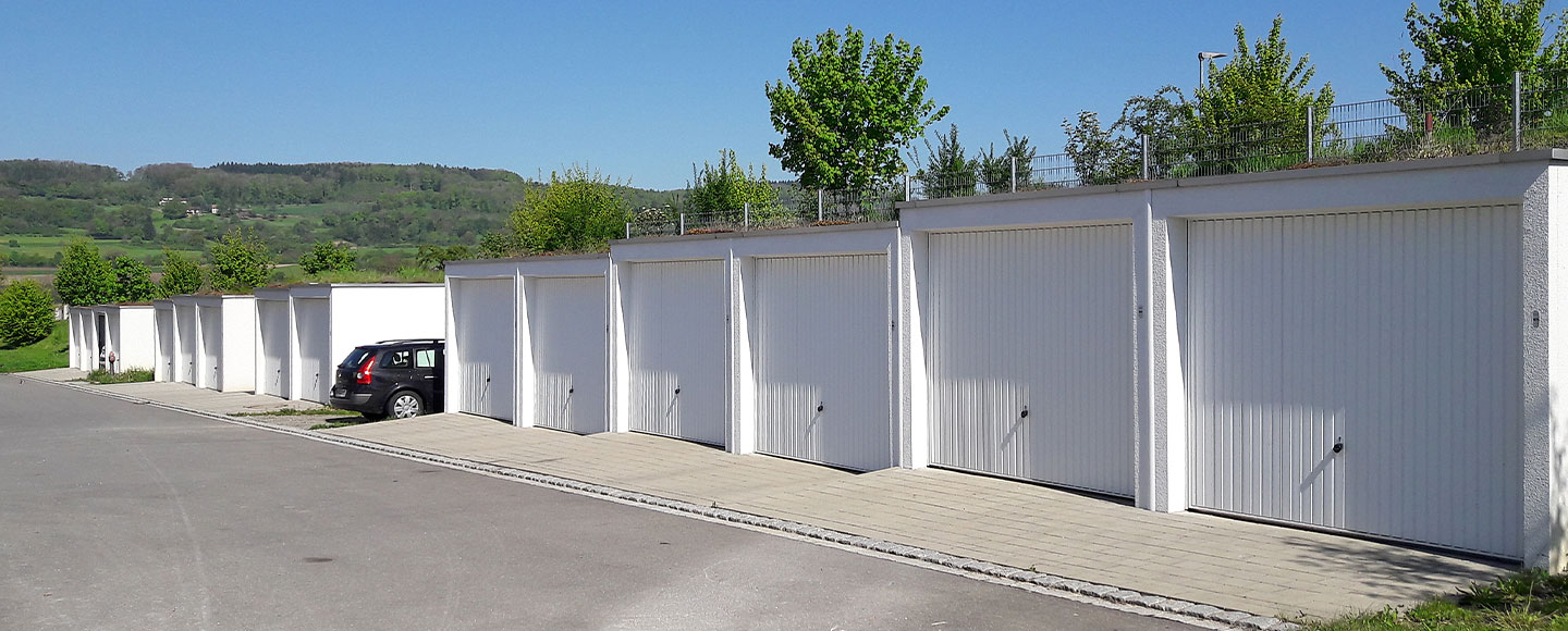 Das Bild zeigt eine Reihe von 14 Garagen, die in einer Treppenoptik angeordnet sind. Nach der sechsten Garage steht ein dunkler Kombi-Pkw geparkt. Im Hintergrund ist eine natürliche Landschaft zu sehen, die einen ruhigen Kontrast zu der strukturierten Garagenreihe bildet.