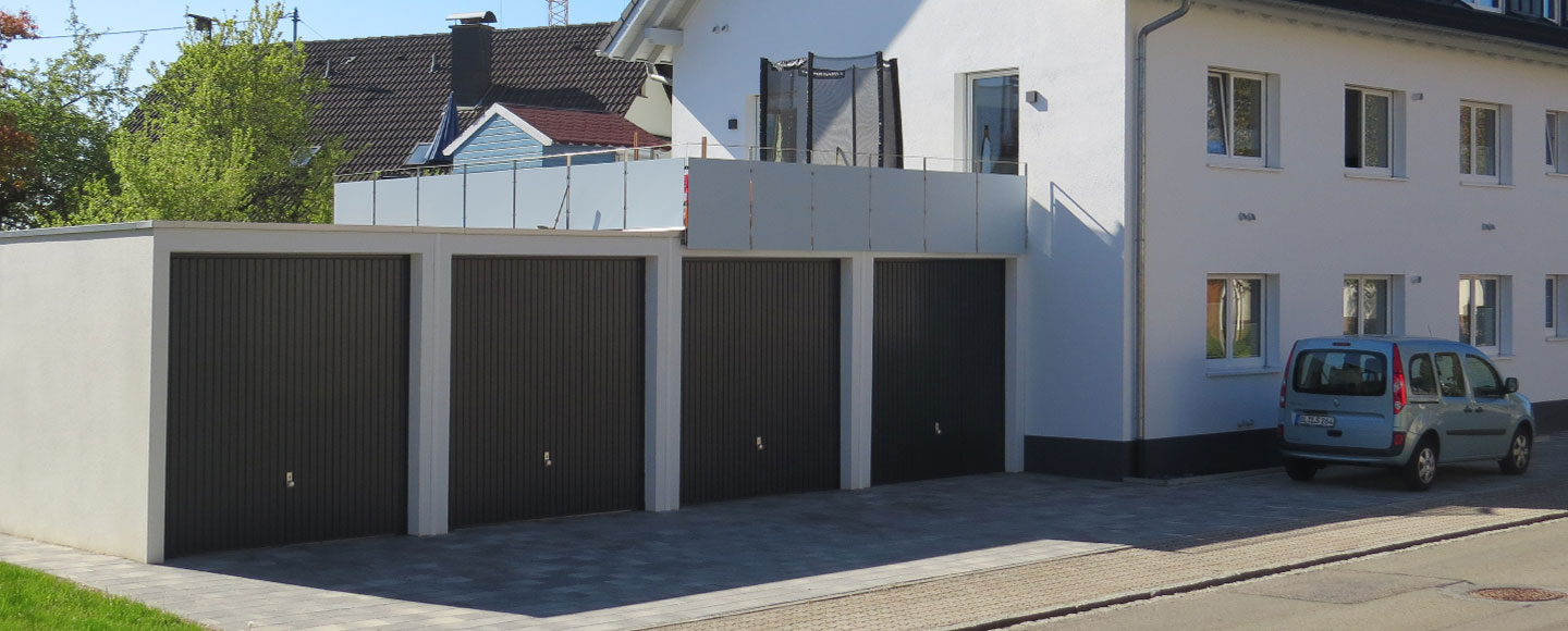 Das Bild zeigt vier aneinandergebaute weiße Einzelgaragen mit braunen Toren. Auf den letzten zwei Garagen wurde eine Dachterrasse mit Metallpanelen als Geländer errichtet, auf der ein Trampolin steht. Rechts angrenzend befindet sich ein weißes Wohnhaus mit einer braunen Einfassung im Erdgeschoss. Vor dem Haus ist ein Auto geparkt