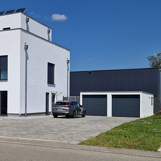 Die Abbildung zeigt eine moderne Doppelgarage mit zwei dunklen Toren. Vor einem Tor ist ein silbernes Auto geparkt. Rechts neben der Garage befindet sich ein Gebäude mit geschlossenen, dunklen Rollläden. Ein schmaler Rasenstreifen trennt die Garage vom angrenzenden Bereich. Im Hintergrund erhebt sich ein schwarzes Gebäude, das die Szene architektonisch einrahmt.
