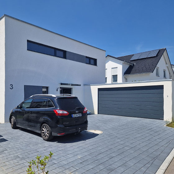 Abgebildet ist eine weiße Großraumgarage, die direkt an ein ebenfalls weißes Wohnhaus angebaut ist. Das Garagentor, die Fensterrahmen und die Türen sind in modernem Anthrazit gehalten. Vor dem Haus mit der Nummer 3 steht ein schwarzes Auto – die Szenerie zeigt eine harmonische Kombination aus Garage und Wohnhaus in zeitgemäßer Architektur.