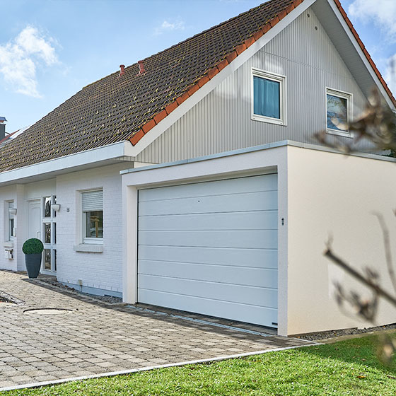 Die Aufnahme zeigt eine weiße Garage neben einem ebenfalls weißen Wohnhaus. Vor dem Haus steht ein großer, hoher Blumentopf mit einem dicht gewachsenen grünen Strauch, der dem Eingangsbereich ein gepflegtes und einladendes Erscheinungsbild verleiht.“