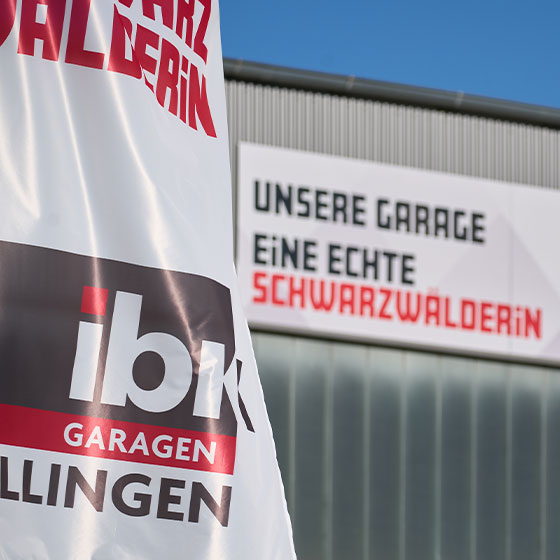 Das Bild zeigt die Fahne von IBK Garagen in Detailaufnahme. Im Hintergrund, sehr verschwommen, wir die Halle gezeigt mit dem Schild "Unsere Garage - Eine echte Schwarzwälderin".