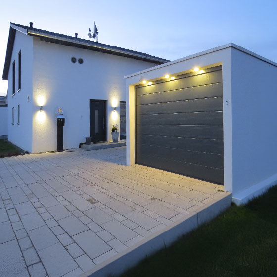 Eine weiße Einzelgarage mit einem anthrazitfarbenen Tor. Das Tor wird von oben beleuchtet. Es ist Dämmerung. Rechts von der Garage befindet sich das Wohnhaus, das ebenfalls beleuchtet ist.