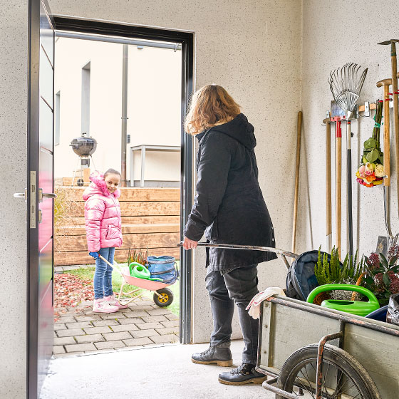 Praktische Nutzräume für mehr Platz - Eine Frau zieht einen kleinen Anhänger mit Gartensachen, darunter eine Gießkanne, in Richtung einer Tür in der Garage. Ihre Tochter wartet draußen mit einer kleinen, rosafarbenen Schubkarre, in der sich ebenfalls eine kleine Gießkanne befindet. Sie trägt eine rosafarbene Jacke mit Fell und rosafarbene Turnschuhe. Draußen liegt Laub. In der Garage hängen weitere Gartengeräte an der Wand.
keywords: Gartenarbeit, Mutter und Tochter, Gießkanne, Schubkarre, Garage, Herbst, Laub, Gartengeräte