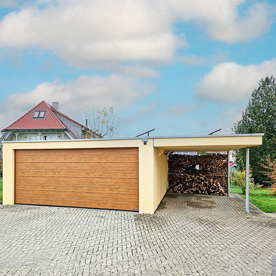 Individuelle Lösungen - Eine Großgarage mit angrenzendem Carport, die zusammen ein harmonisches Bild abgeben. Die Garage ist hellgelb gestrichen, das Holztor passt perfekt dazu. Im hinteren Teil des Carports ist Feuerholz gestapelt.