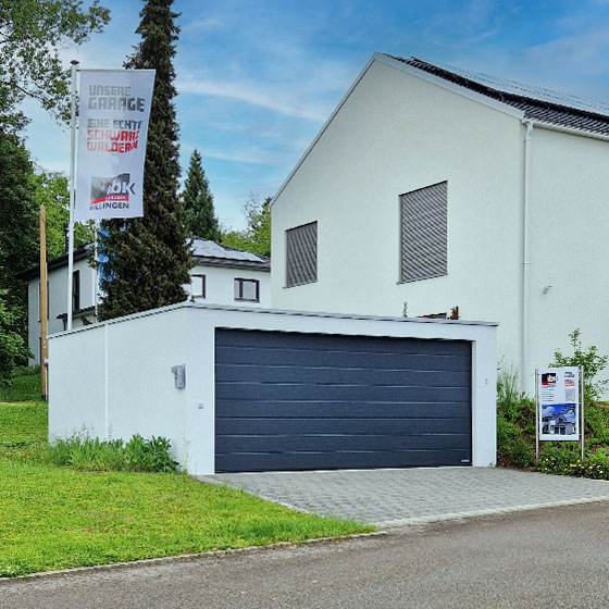 Zu sehen ist eine Großgarage mit dunklem Tor von IBK Garagen Villingen, ausgestellt im HausBauPark Schwenningen. Hinter der Garage weht eine IBK-Fahne, rechts daneben steht ein Informationsschild mit weiteren Details. Im Hintergrund befindet sich ein weißes Wohnhaus mit geschlossenen Fensterrollläden.