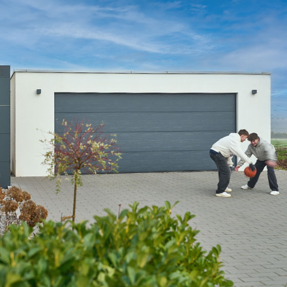 Das Bild zeigt eine moderne Duo-Garage in Weiß mit einem anthrazitfarbenen Garagentor. Vor der Garage spielen zwei Jungen Basketball, was der Szene eine lebendige und familienfreundliche Atmosphäre verleiht.
