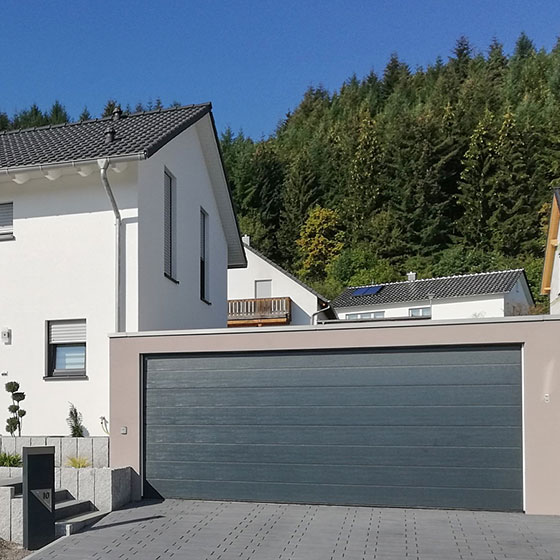 Das Bild zeigt eine beigefarbene Großraumgarage mit einem durchgehenden, anthrazitfarbenen Garagentor. Im Hintergrund ist das Wohnhaus sichtbar. Ein paar steinerne Stufen führen zum Hauseingang mit einer Tür in Anthrazit.