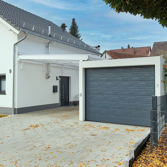Das Bild zeigt eine weiße Einzelgarage mit grauem Garagentor. Ein modernes Glasdach verbindet die Garage mit dem angrenzenden Wohnhaus. Unter dem Glasdach befindet sich die Haustür in einem eleganten Anthrazitton.