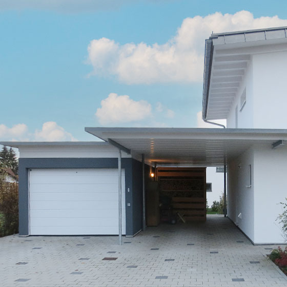 Abgebildet ist eine Garage des Modells Grando in anthrazitfarbener Ausführung. Das weiße Garagentor bildet einen markanten Kontrast. Oberhalb der Garage befindet sich eine weiße Holzerhöhung, die in eine Überdachung übergeht. Diese Überdachung schafft eine optische und funktionale Verbindung zum angrenzenden Wohnhaus.