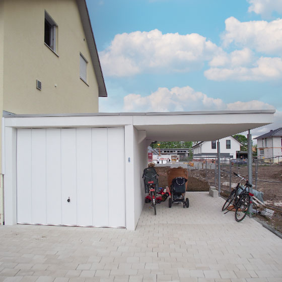 Das Bild zeigt eine weiße Kombination aus Garage und Carport, getragen von metallenen Säulen. Unter dem offenen Carport-Bereich stehen ein Fahrrad mit Kindersitz, ein Kinderwagen und ein normales Fahrrad. Rechts neben der Garagenanlage befindet sich eine Baustelle, links ist ein Wohnhaus in Eierschalenfarbe zu sehen