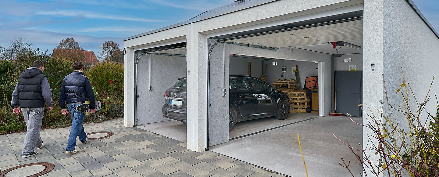 Zu sehen ist eine offene Doppelgarage mit einem Durchbruch in der Mitte, der dem Nutzer zusätzlichen Platz bietet. Auf der linken Seite der Garage steht ein schwarzes Auto, während die rechte Seite leer ist. Vor der Garage laufen zwei Teenager vorbei – einer trägt ein Skateboard unter dem rechten Arm. Beide sind von hinten zu sehen und tragen Jeans. Die Szene vermittelt einen modernen, jugendlichen Alltagseindruck.