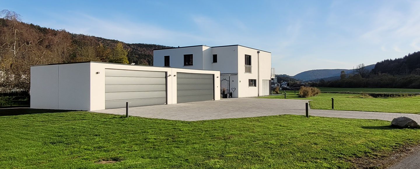 Das Bild zeigt zwei nebeneinander gebaute Großraumgaragen in Weiß mit jeweils einem dunklen Garagentor. Direkt daneben befindet sich ein modern gestaltetes, weißes Flachdachhaus mit dunklen Fensterrahmen. Das gesamte Ensemble – Garagen und Wohnhaus – steht auf einem weitläufigen Rasenstück und vermittelt ein einheitliches, modernes Erscheinungsbild.