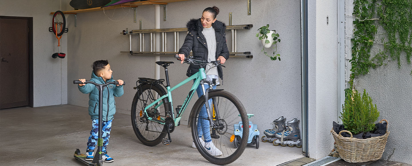 Das Bild zeigt eine Alltagsszene: Eine Mutter mit einem Fahrrad und ihr Sohn mit einem Roller kommen gemeinsam aus einer weißen Großraumgarage. Im Inneren der Garage ist eine Leiter an der Wand befestigt. Die Szene vermittelt Familienfreundlichkeit, Funktionalität und Ordnung im Garagenalltag