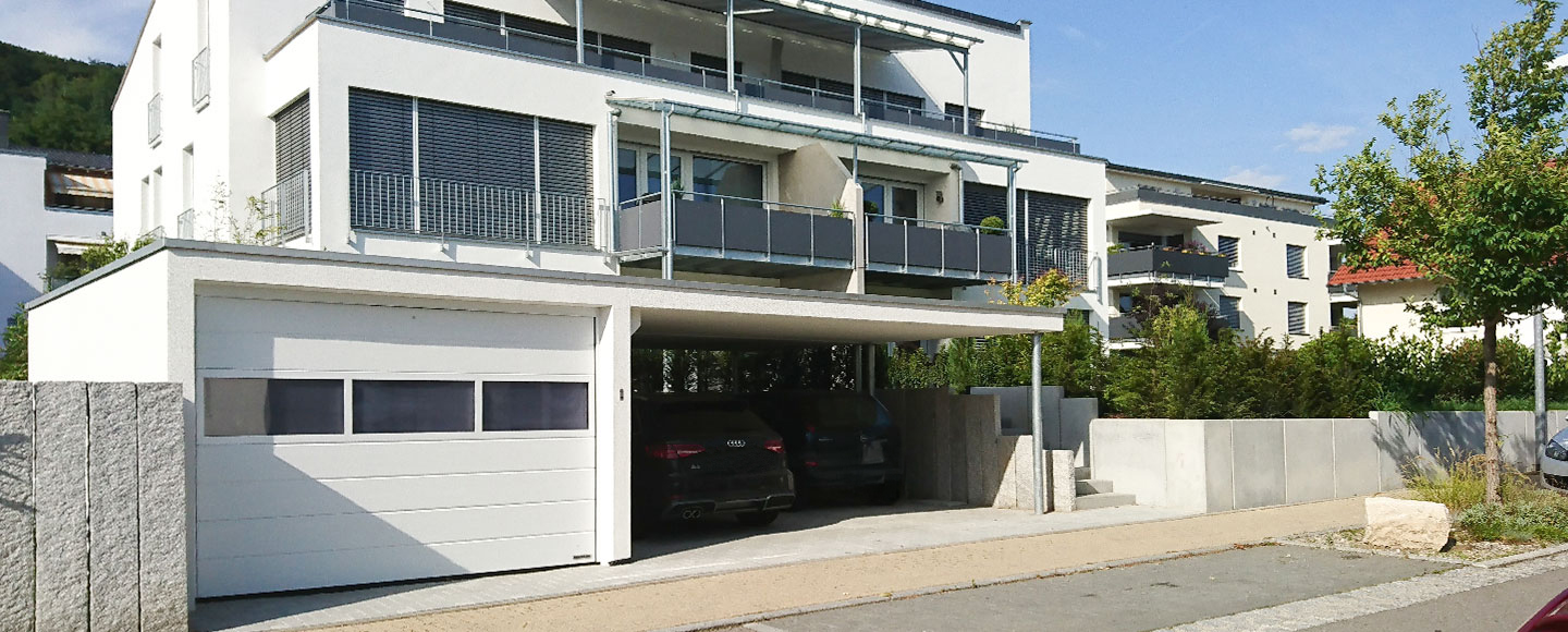 Das Bild zeigt eine moderne Garagen-Carport-Kombination, die in die Betonumzäunung eines Mehrfamilienhauses integriert ist. Die Umzäunung rahmt sowohl den Garten als auch das erhöht gebaute Wohnhaus ein. Der Garten ist mit dicht wachsenden Tujabüschen bepflanzt, die als Sichtschutz dienen. Die Garage verfügt über eine dreiteilige Fensterfront. Im Carport stehen zwei schwarze Autos nebeneinander.