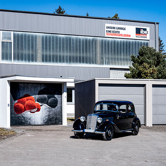 Auf dem Hof von IBK Garagen in Villingen stehen eine Einzel- und eine Doppelgarage. Die Einzelgarage ist mit traditionellen roten und schwarzen Bollenhüten aus dem Schwarzwald bemalt. Schräg davor ist ein schwarzer Oldtimer geparkt, der den regionalen Charakter der Szene unterstreicht