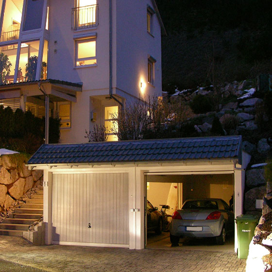 Das Bild zeigt eine Doppelgarage mit Giebeldach. Es ist schon Nacht und die Lichter im Innern der Garage wie auch die Lichter im Haus sind an. Die Garage liegt am Hang. Ein Garagentor ist geschlossen und ein Garagentor ist offen und ein Silberner Kleinwagen ist ersichtlich. Links von der Garage gehen Stufen nach oben zum Wohnhaus.