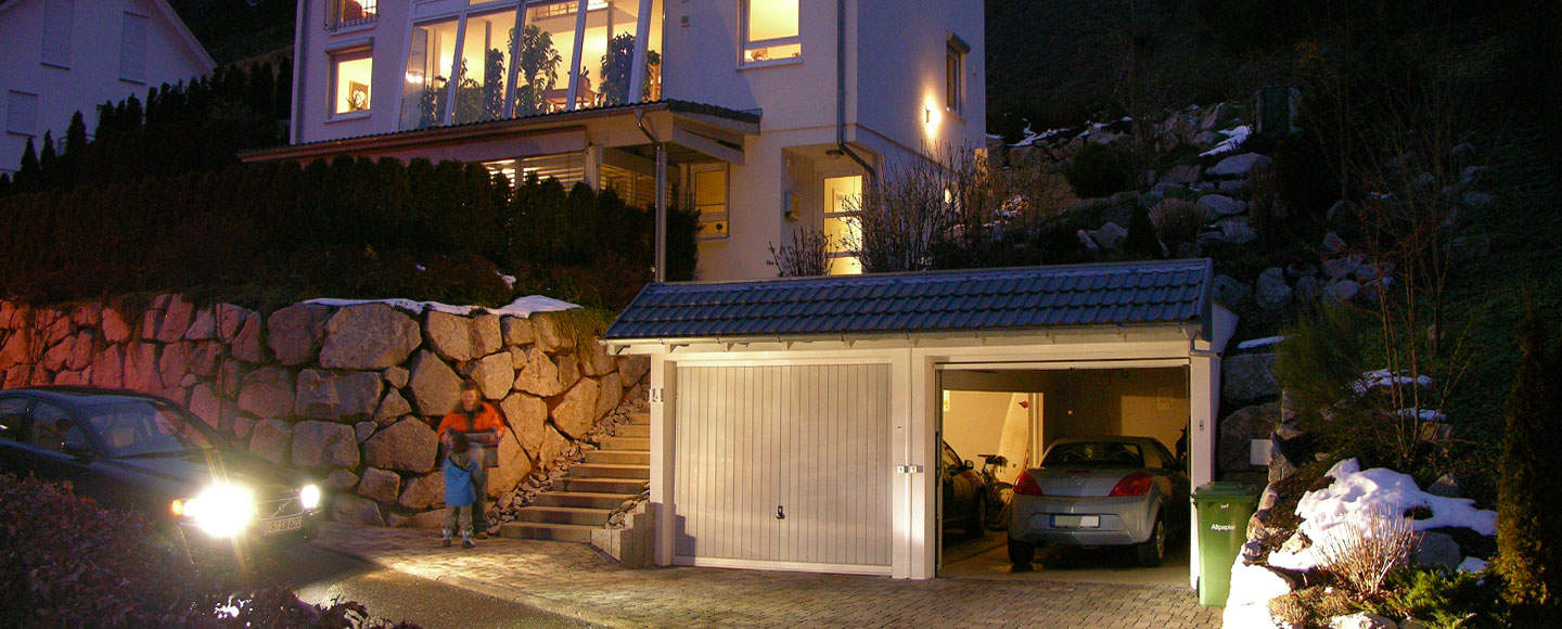 Bewährte Handarbeit und moderne Technik - Eine Doppelgarage mit Giebeldach, die am Hang liegt. Es ist Nacht, und sowohl die Lichter in der Garage als auch im Wohnhaus sind eingeschaltet.