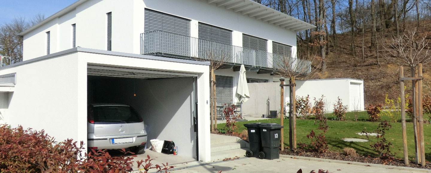 Das Bild zeigt eine Einzelgarage in der ein Auto steht im Vordergrund. Daneben steht zwei Mülltonnen. Rechts hinter der Garage steht ein Mehrfamilienhaus mit herunter gelassenen Rollläden. und einem riesigen, über die gesamte breite des Hauses gehender Balkon aus Metall. Rechst neben dem Haus steht nochmal eine Garage in weiß.